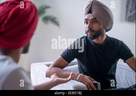 Indian man wearing turban looking at his friend talking sitting on sofa at home Stock Photo