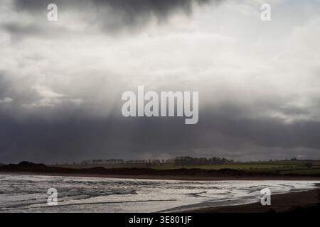 Sunbeams pierce through dark, moody storm clouds over a serene beach and rolling green hills, evoking a sense of nature's power and beauty Stock Photo