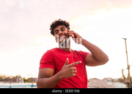 Cheerful Arab man wearing red shirt having phone call while standing outdoors Stock Photo