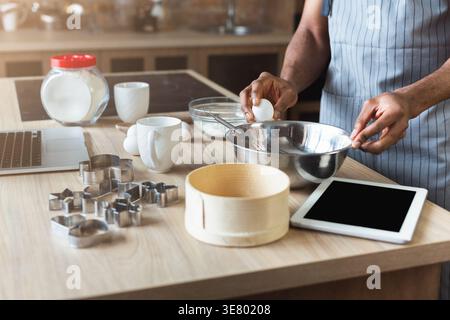 Black man in apron baking while standing at table with laptop in kitchen at home Stock Photo
