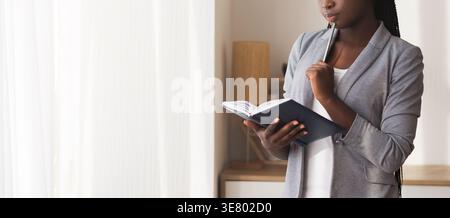 Cropped image of Black woman with twists holding planner and pen on chin standing in the office during daytime Stock Photo