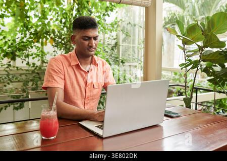 Indian male entrepreneur working on laptop sitting at table with drinks in cafe Stock Photo
