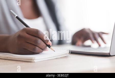 Closeup of Black woman typing on laptop and writing in notebook sitting at table in the office during daytime Stock Photo