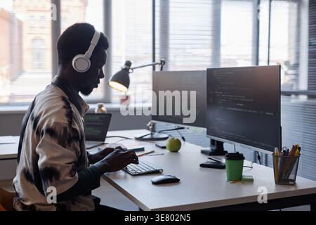 Black male programmer wearing headphones typing codes on computer while sitting at table in office Stock Photo