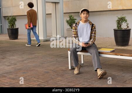 Diverse male students sitting on campus bench holding phone, walking away with folders, copy space Stock Photo