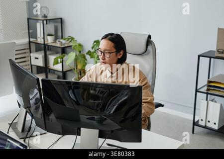 Serious Asian male programmer wearing eyeglasses working on laptop sitting at table in office Stock Photo