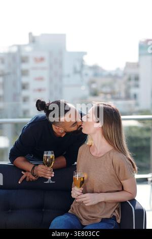 Middle Eastern man with top knot kissing his caucasian girlfriend holding wine glass while sitting on sofa in balcony at home Stock Photo
