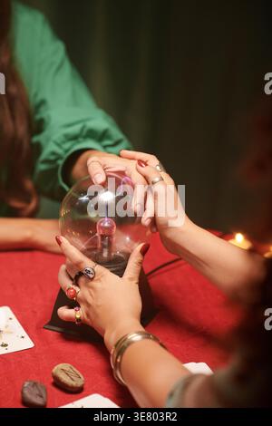 Mixed-race female fortune teller using crystal ball with caucasian female client while sitting at table with rocks and tarot cards indoors Stock Photo