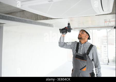 Indian male worker wearing uniform and hard hat cleaning and repairing air conditioner in an empty building Stock Photo