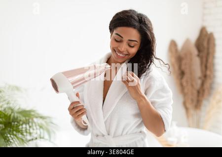 Black woman in bathrobe blow drying hair standing in bathroom at home Stock Photo
