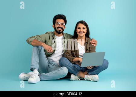 Smiling Asian couple with laptop sitting with crossed legs on floor against blue background in studio Stock Photo