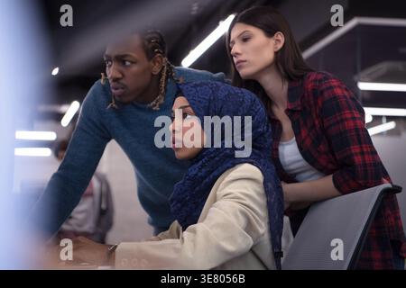 Black business woman with hijab sitting at table working on computer with coworkers standing beside her in office Stock Photo