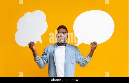 Confused Black man holding two speech bubbles standing against yellow background in studio Stock Photo