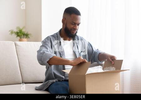 Black man unpacking cardboard box while sitting on sofa in living room at home Stock Photo