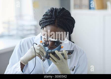 Focused Black female computer engineer with braids wearing magnifying glasses examining electronic parts sitting at table in engineering laboratory Stock Photo