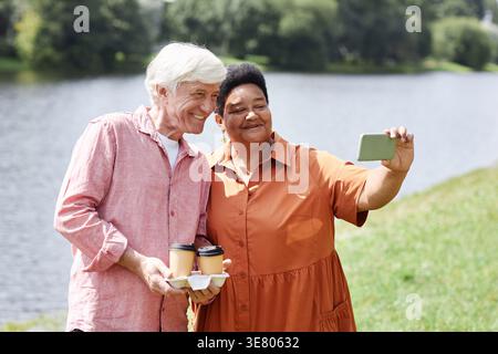 Black middle-aged woman holding smartphone taking selfies with caucasian senior man standing near the lake Stock Photo