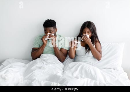 Black couple having fever measuring body temperature with thermometer blowing nose using napkin sitting on bed at home Stock Photo