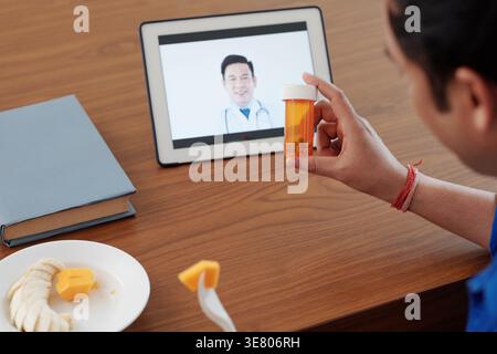 Indian man holding bottle of medicine having online consultation with Asian doctor on tablet sitting at table at home Stock Photo