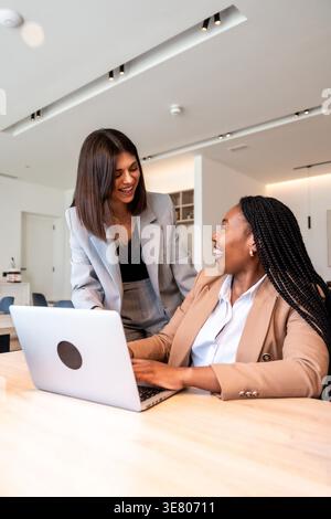 Black and caucasian businesswomen talking with each other and working on laptop while standing and sitting at table in office Stock Photo