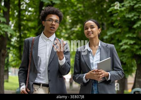 Black business woman holding tablet and looking away while walking beside male colleague wearing eyeglasses discussing project details in park duri... Stock Photo