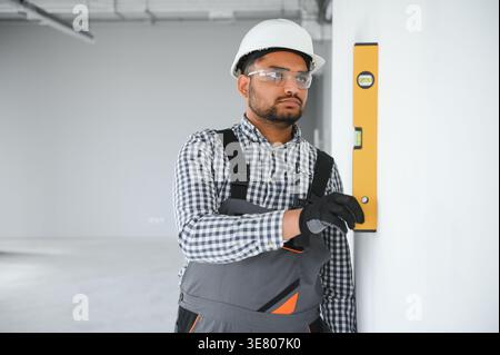 Indian male builder wearing uniform and hard hat using water level measuring device during work in an apartment building Stock Photo