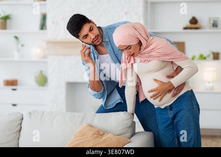 Worried Middle Eastern husband having phone call and supporting pregnant wife suffering prenatal contractions while walking in living room at home Stock Photo