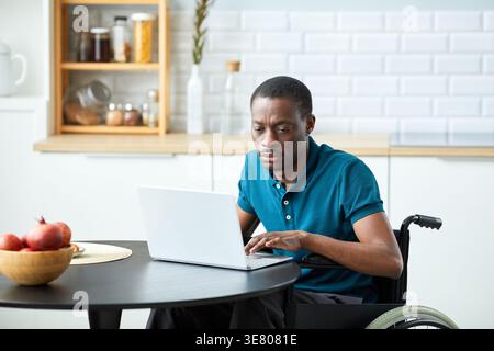 Disabled Black man in wheelchair working on laptop at table in kitchen at home Stock Photo