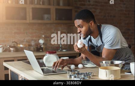 Black man in apron holding eggs and using laptop while standing in kitchen at home Stock Photo