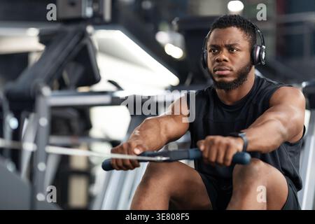 Focused Black male listening to music with headphones working out using rowing machine in gym during workout Stock Photo