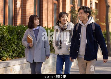 Smiling Chinese female college students walking and talking with male classmate on campus on sunny day Stock Photo