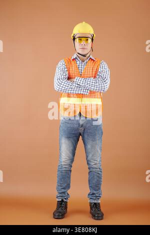 Asian male civil engineer wearing hard hat safety goggles safety reflective vest and safety shoes with arms crossed to chest standing against brown... Stock Photo