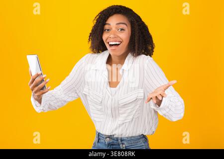 Surprised Black woman with curly hair shoulders up arms slightly open holding phone and making hand gesture standing against yellow background in s... Stock Photo