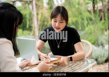 Asian woman working remotely with her colleague while sitting at table in garden during daytime Stock Photo