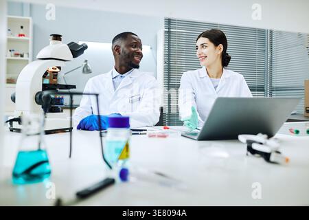 Smiling Black and mixed-race scientists wearing lab coats and protective gloves talking with each other while sitting together at table with laptop... Stock Photo