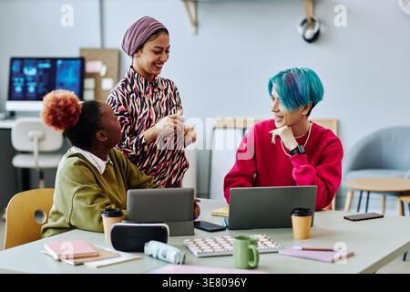 Asian male programmer talking with Black and mixed-race female colleagues while sitting at table with laptop and tablet in office Stock Photo