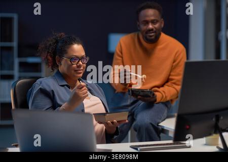 Black male and mixed-race female programmers eating dinner together discussing computer program while working late at night in office Stock Photo