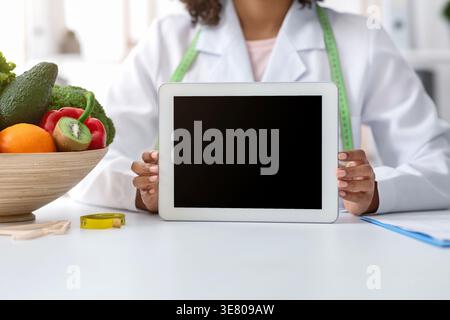 Cropped image of Black female nutritionist with tape measure around her neck holding tablet with blank screen while sitting at table with bowl of f... Stock Photo
