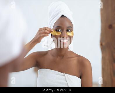 Black woman wrapped in bath towel after shower applying anti-fatigue under eye mask standing in front of mirror in bedroom at home Stock Photo
