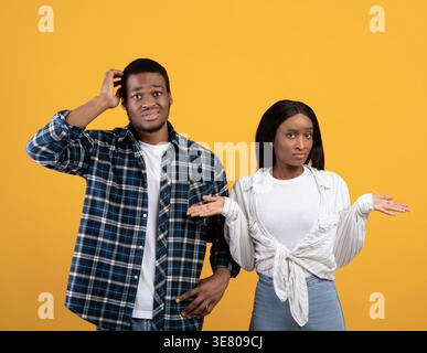 Confused Black couple shrugging shoulders and touching head while standing against yellow background in studio Stock Photo