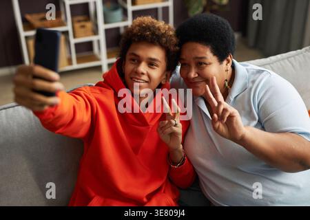 Smiling Black teenage son with mother gesturing peace sign taking selfie using smartphone while sitting on sofa in living room at home Stock Photo