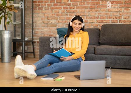 Middle Eastern female student with headphones writing down on notebook looking at laptop while sitting on floor near sofa in living room at home Stock Photo