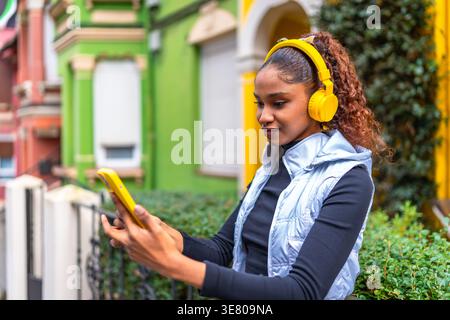 Cheerful Hispanic woman with curly hair listening to music on headphones and browsing on smLifestylephone while standing in city during daytime Stock Photo