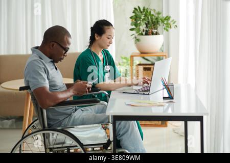 Asian female social worker working on laptop beside disabled Black male patient using smartphone sitting in wheelchair at home Stock Photo