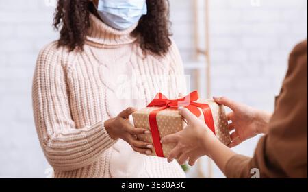 Black woman with curly hair wearing medical mask receiving gift from female friend while standing indoors during pandemic Stock Photo