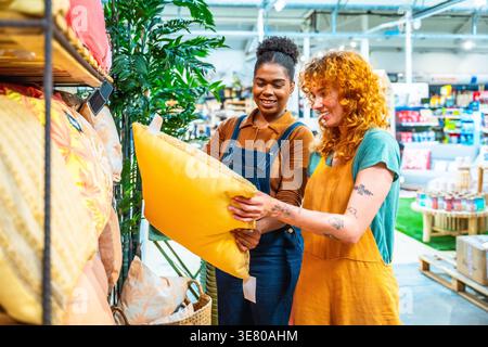 Hispanic and caucasian female friends holding and looking at yellow pillow while standing together in home furnishing store Stock Photo