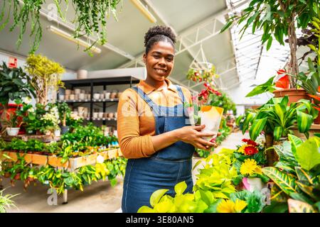 Hispanic female gardener in denim overalls holding potted plant and smiling while standing in greenhouse Stock Photo
