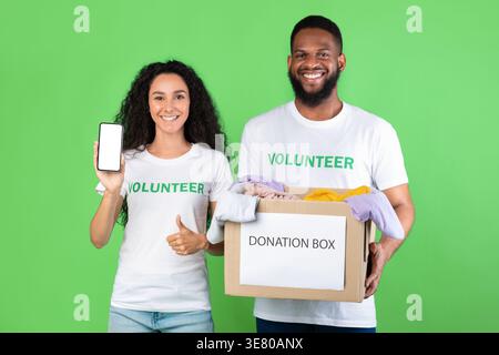 Smiling diverse group of volunteers showing smartphone and holding donation box standing against green background in studio Stock Photo