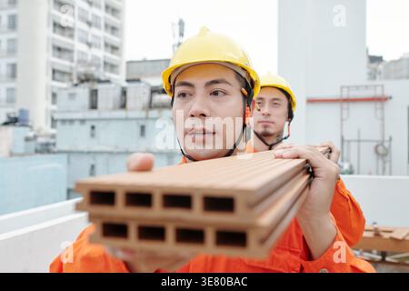 Asian and mixed race male construction workers carrying wood flooring boards while standing in construction site Stock Photo