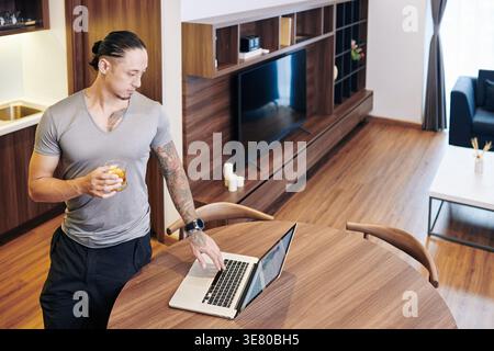 Mixed-race man holding glass of whiskey with slice of lemon checking graph on his laptop standing at table in apartment Stock Photo