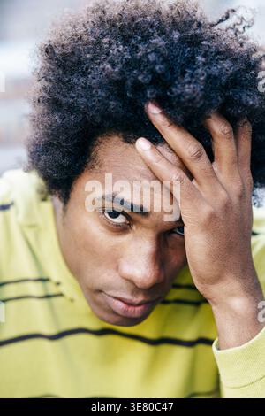 Serious Black man with afro hair sitting on ground with hand on head looking away outdoors Stock Photo